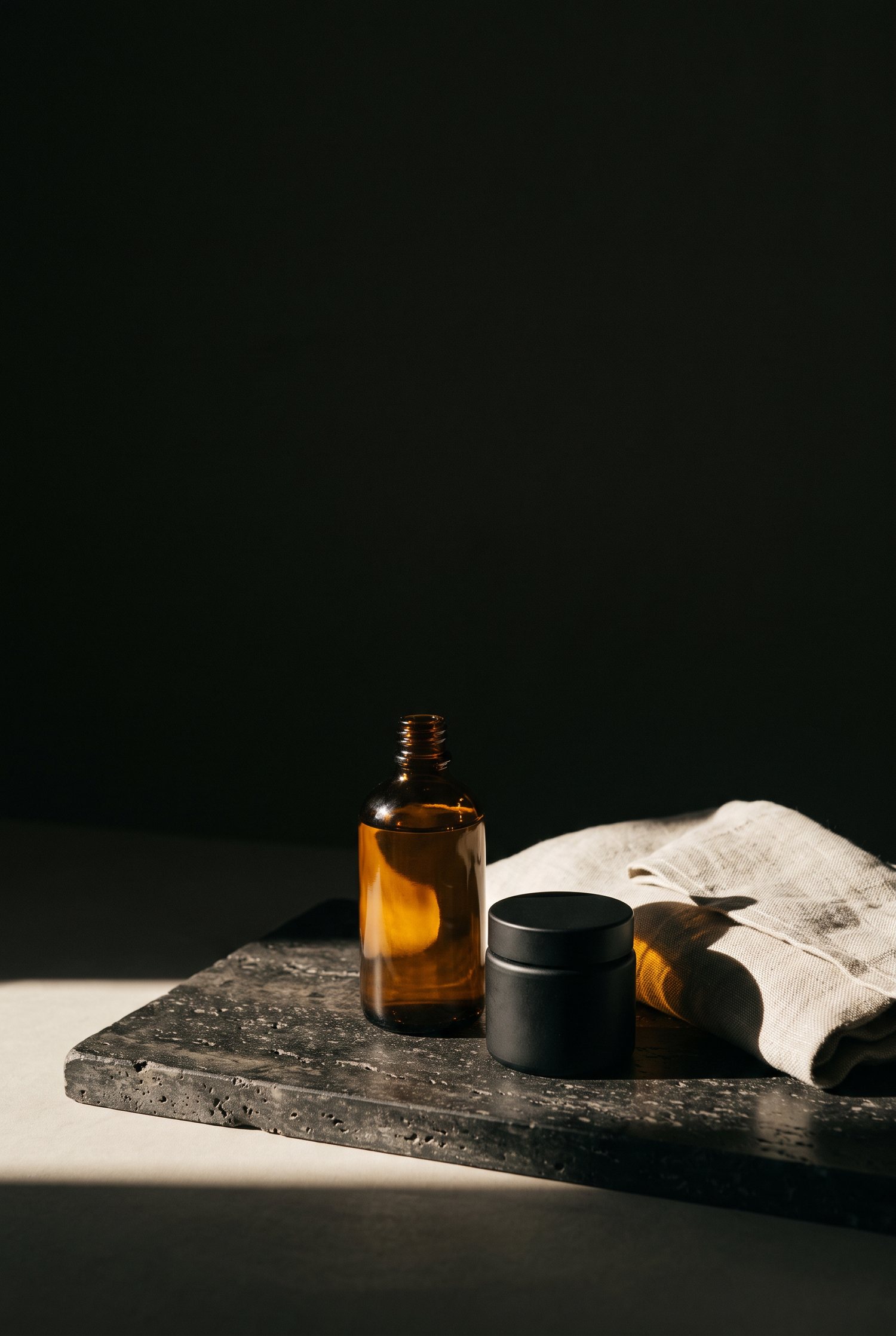 An amber glass bottle and a black jar on a dark stone slab beside folded linen, lit by low sunlight.